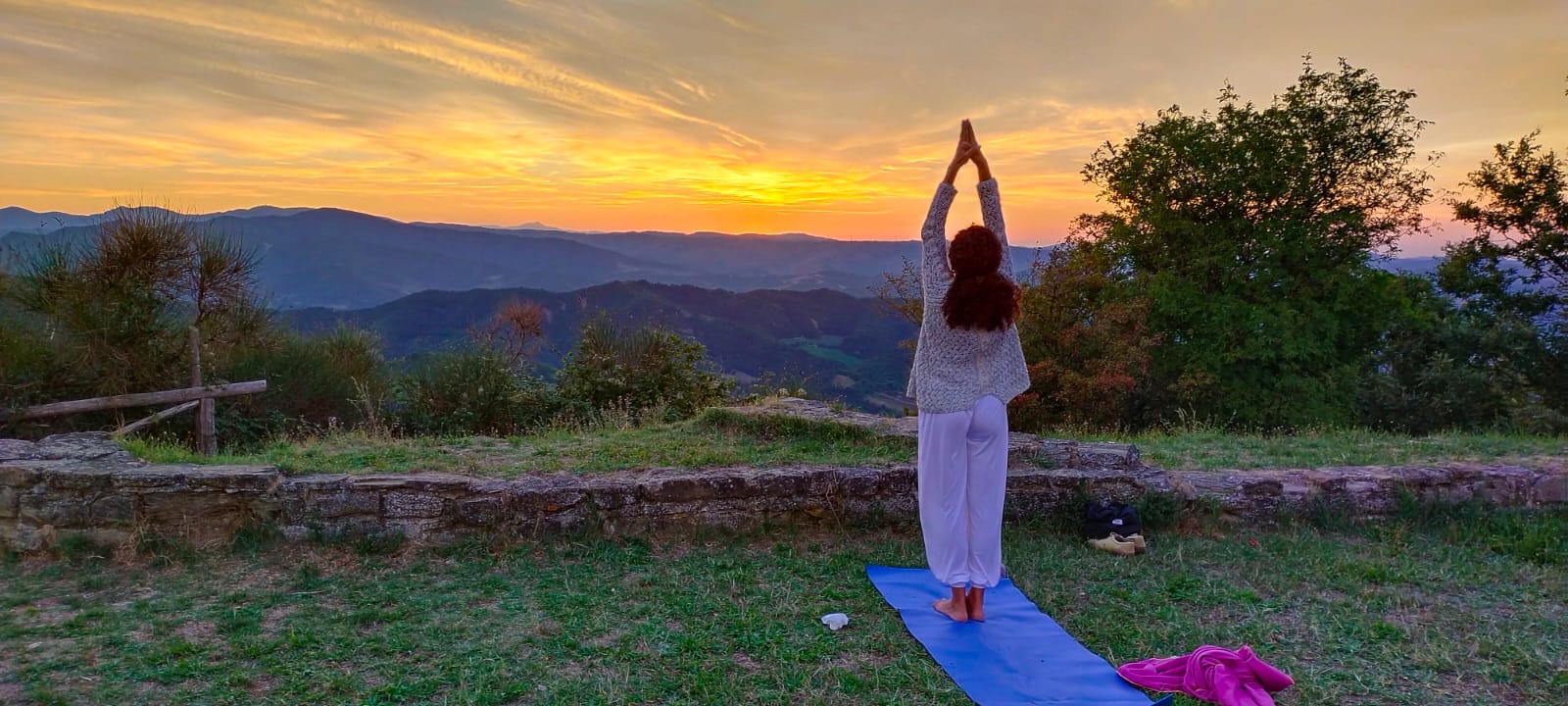 A person practicing yoga at sunset on a blue mat, surrounded by mountains and greenery.