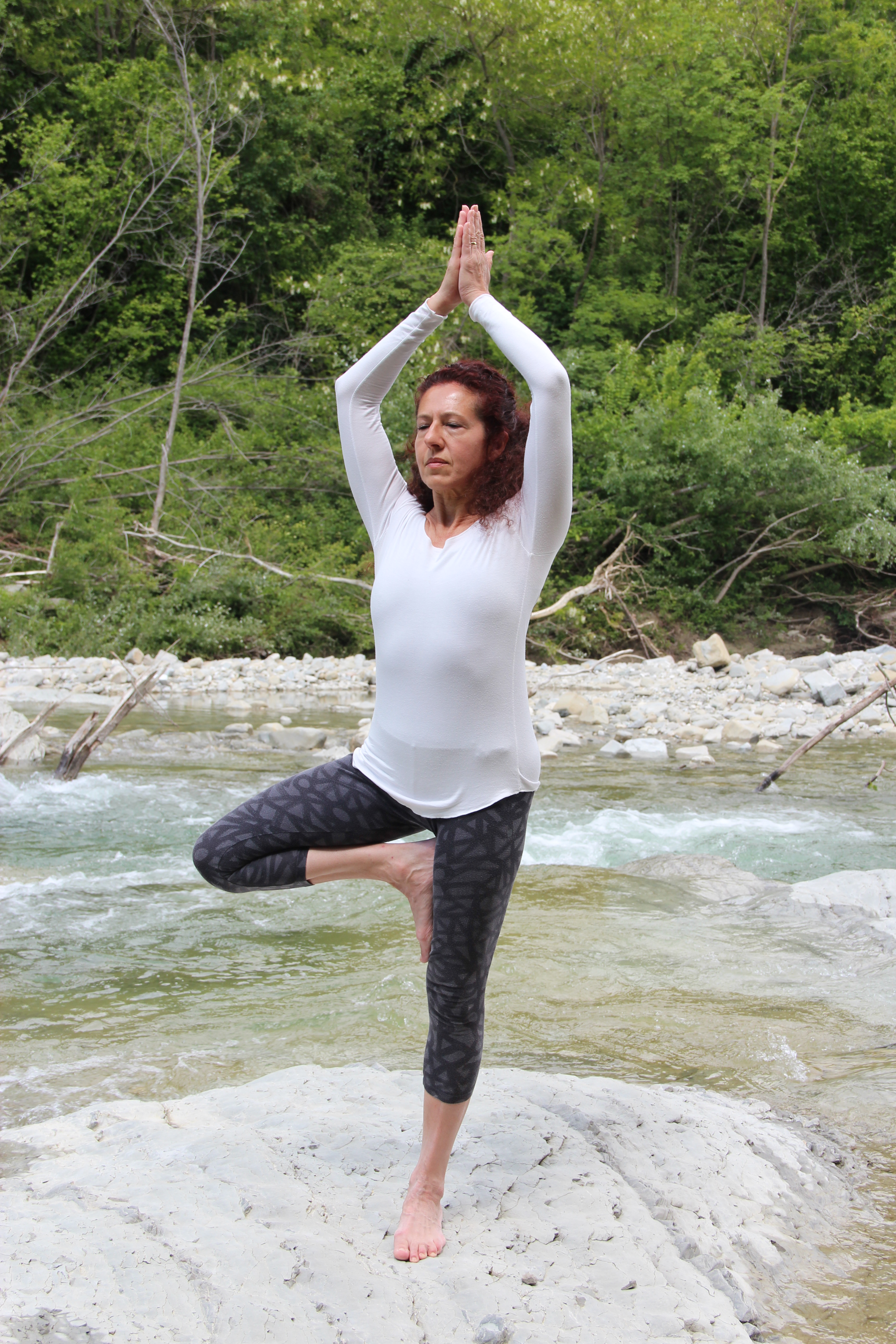 A woman performing a yoga pose on one leg while standing on a rock near a river, surrounded by lush greenery.
